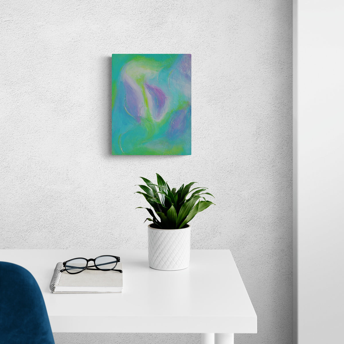 An abstract canvas painting with swirls of teal, green, and purple hangs on a light gray textured wall above a white desk with a potted plant and glasses.